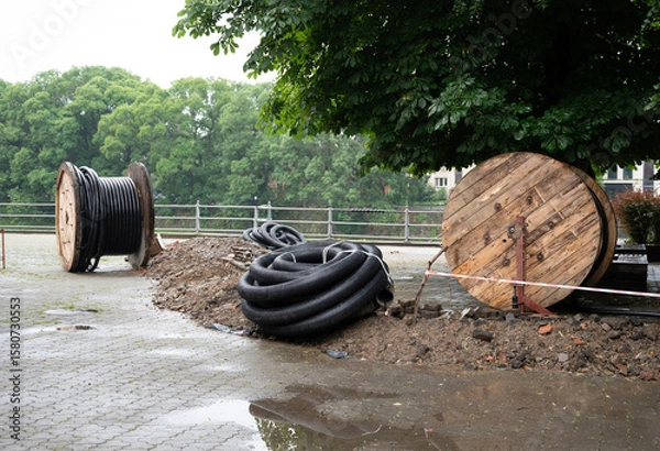 Fototapeta Coiled cables and wooden spools near a green park area with trees during a cloudy day