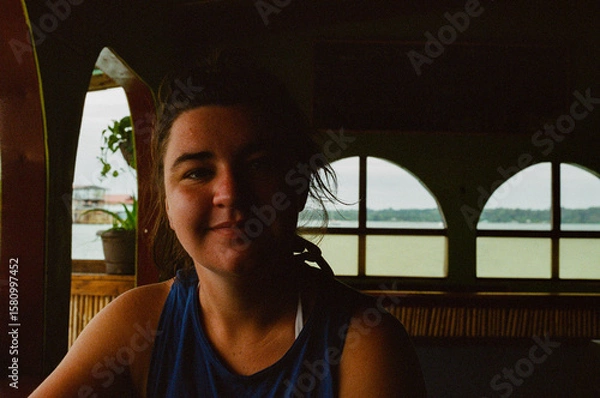 Fototapeta A woman seated in a cozy oceanside interior with natural lighting, looking relaxed and thoughtful against the backdrop of the calm ocean seen through large windows adorned with curved frames.35mm film
