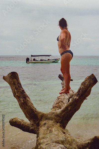 Fototapeta A woman on a tropical beach stands on a driftwood log overlooking serene turquoise waters, with a boat anchored in the distance, evoking a sense of adventure, relaxation, and scenic beauty. 35mm film
