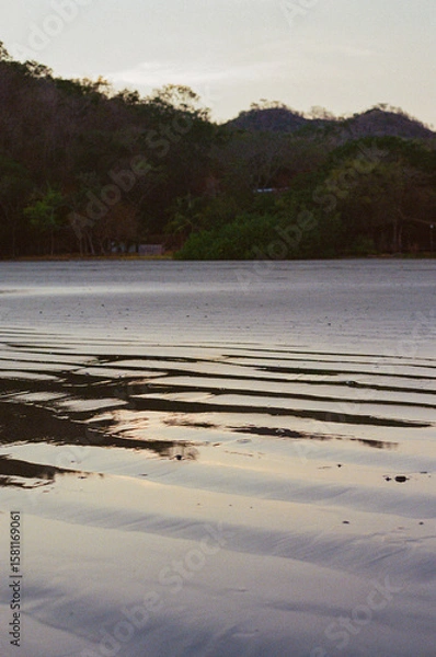 Fototapeta An idyllic beach scene showing rippling sands under soft twilight reflections, surrounded by lush foliage and distant hills, evoking serenity and calm with its peaceful ambiance. Taken on 35mm film