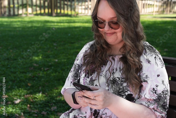 Fototapeta Young woman with long curly hair using a smartphone outdoors in a park on a sunny day, smiling and relaxed.