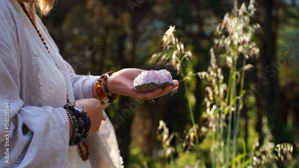 Fototapeta A woman's hand in a white kimono holds a pink crystal in the mountains of Sintra. Close-Up.  Soft focus