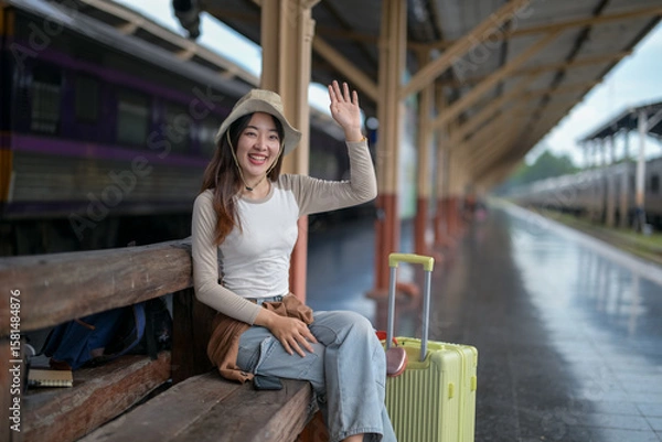 Fototapeta A woman is sitting on a bench at a train station with a suitcase in front of her