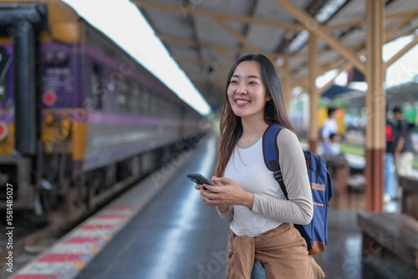 Fototapeta A woman is smiling and holding a cell phone while standing at a train station
