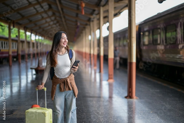 Fototapeta A woman is walking through a train station with a suitcase and a cell phone