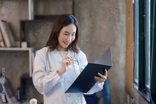 Fototapeta Asian cheerful happy female professional successful doctor practitioner in a white lab coat with a stethoscope standing smiling holding a reading paperwork document in a folder in the hospital.