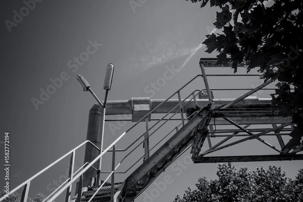 Fototapeta Industrial staircase leads to the sky above a tree line on a bright day, showcasing a blend of nature and urban structures