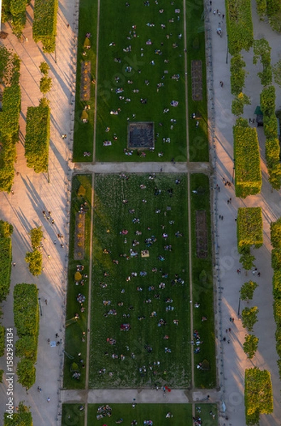 Fototapeta An aerial view from above of people relaxing and picnicking on the green lawns of the Champ de Mars in Paris, France, at sunset, taking advantage of the last rays of the sun.