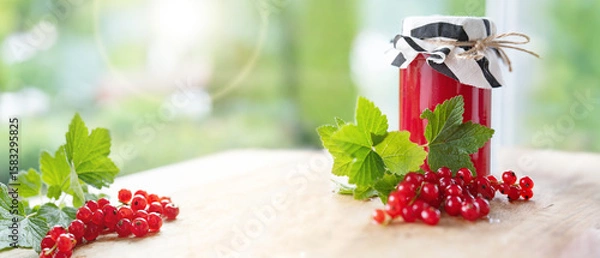 Obraz Fresh homemade red currant jam in jar with currants and green leaves on wooden table against light bokeh background. Horizontal close-up with space for text.