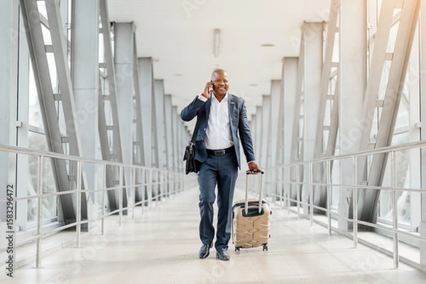 Fototapeta Smiling African American businessman talking on cellphone while walking confidently through a modern airport terminal, black male pulling suitcase and carrying a leather briefcase