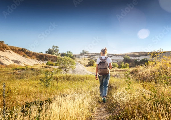 Fototapeta The girl with a backpack walks the field in the rays of the setting sun. Beautiful landscape, freedom, the unity of human and nature