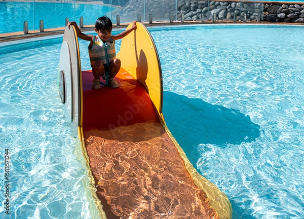 Fototapeta A child is playing in a pool with a slide. The pool is surrounded by trees and has a blue sky above it