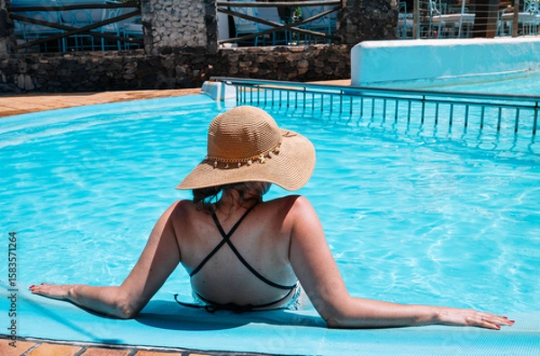 Fototapeta A woman in a black bikini is sitting in a pool with a straw hat on. The pool is blue and the woman is looking at the water
