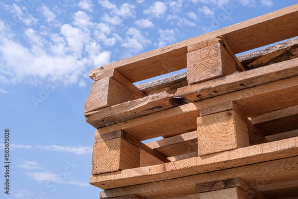 Fototapeta Close-up view of several rough wooden pallets stacked outdoors under a bright blue sky with scattered clouds. Highlighting rustic industrial design and recycling themes.