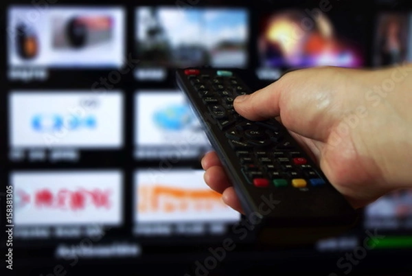 Fototapeta Smart tv and hand pressing remote control.Hand holding TV remote control with a television in the background. Close up.