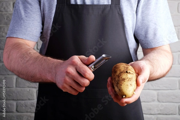 Fototapeta man in black apron holding potatoes and knife for peeling in his hands, on background of brick wall