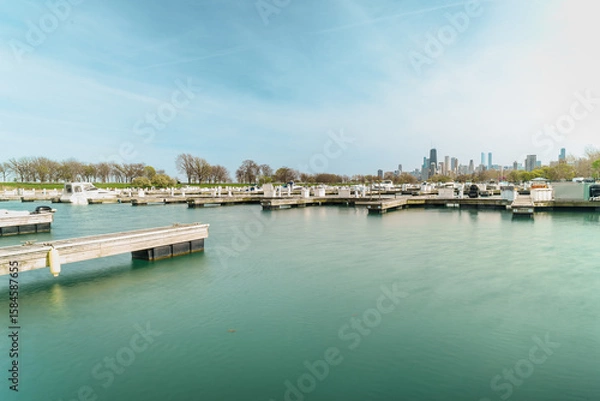 Fototapeta Photograph of a serene marina with numerous docks, calm water reflecting a clear sky, surrounded by bare trees, with a city skyline visible in the background.
