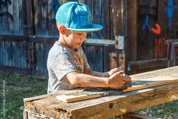 Fototapeta Little boy wearing a cap is focused on shaping clay in the yard of a rustic village house, enjoying a sunny day outdoors