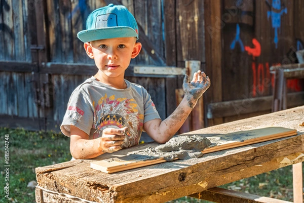 Fototapeta Little boy wearing a cap is focused on shaping clay in the yard of a rustic village house, enjoying a sunny day outdoors
