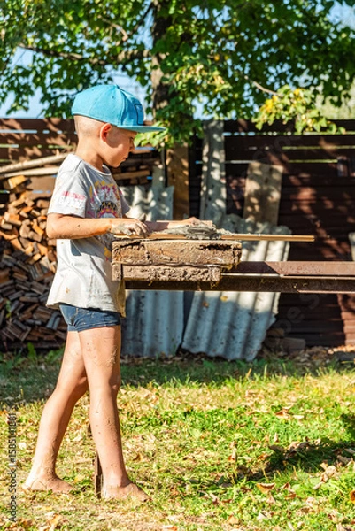 Fototapeta Young boy in a cap cheerfully creates with clay, standing barefoot and covered in dirt, near a rustic wooden yard table