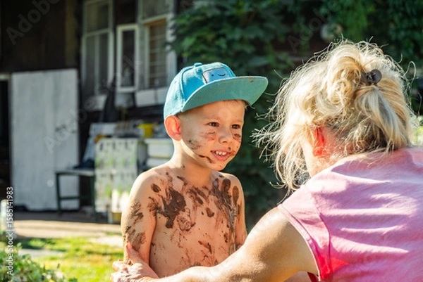 Fototapeta Little boy, dirty and smiling, faces his grandmother who appears to be scolding him in a country yard, on a sunny day