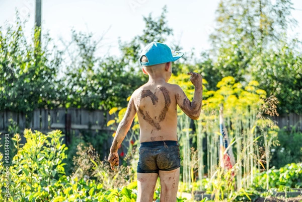 Fototapeta Little boy wearing a cap stands in a garden, covered in mud and proudly displaying a heart shape on his back while having fun outdoors