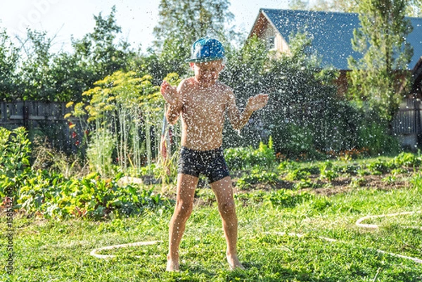 Fototapeta Joyful boy in a cap enjoys splashing water from a hose, playfully cleaning himself in a vibrant garden under bright sunshine