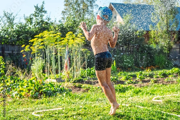 Fototapeta Joyful boy in a cap enjoys splashing water from a hose, playfully cleaning himself in a vibrant garden under bright sunshine