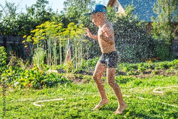 Fototapeta Joyful boy in a cap enjoys splashing water from a hose, playfully cleaning himself in a vibrant garden under bright sunshine