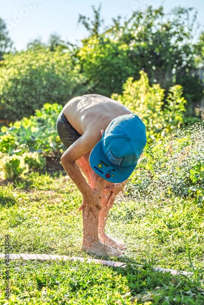 Fototapeta Joyful boy in a cap enjoys splashing water from a hose, playfully cleaning himself in a vibrant garden under bright sunshine