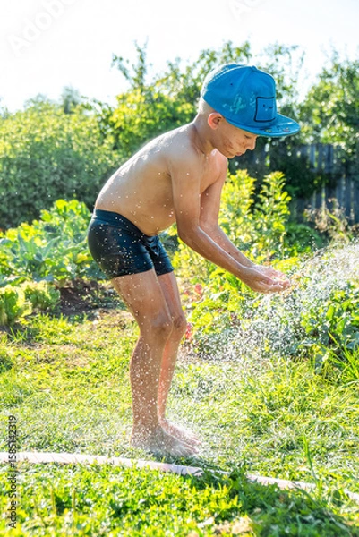 Fototapeta Joyful boy in a cap enjoys splashing water from a hose, playfully cleaning himself in a vibrant garden under bright sunshine