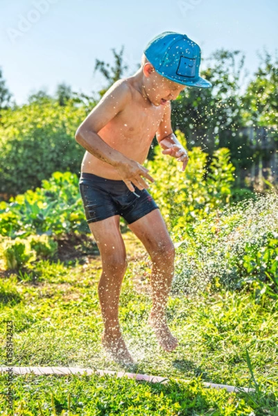 Fototapeta Joyful boy in a cap enjoys splashing water from a hose, playfully cleaning himself in a vibrant garden under bright sunshine