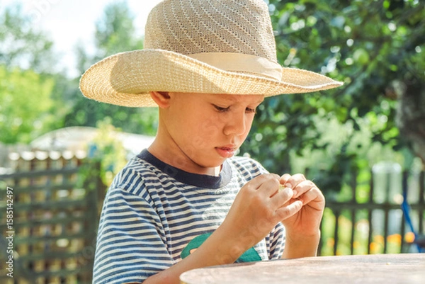 Fototapeta Small boy in a straw hat focuses on shelling beans while sitting at a table outside, surrounded by greenery
