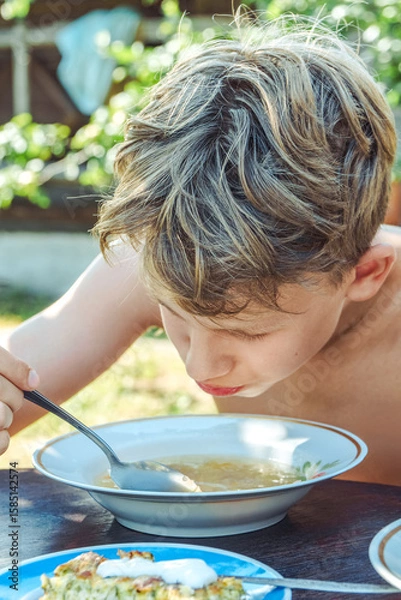Fototapeta Boy happily eats his lunch from a bowl while sitting at a wooden table in a bright, sunny garden of a countryside home, closeup