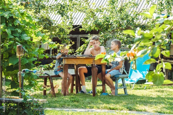 Fototapeta Three brothers are having lunch at a round wooden table in a lush garden, surrounded by greenery and sunlight