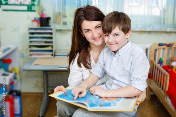 Fototapeta Beautiful happy mother with child boy 10 years old reading book, sitting on chair in children's room at home