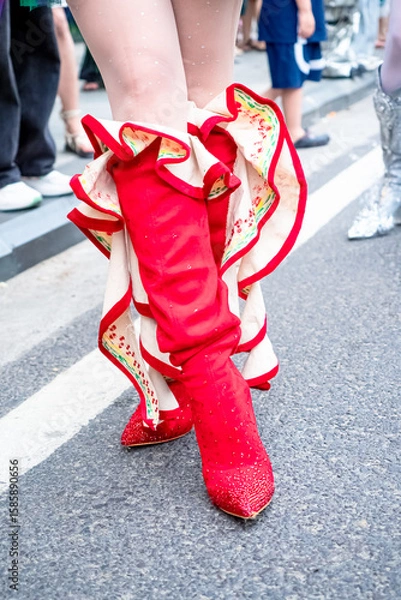 Fototapeta Red carnival boots on women's legs close-up against the background of a street carnival procession: bright costume details, festive atmosphere, dancing, street, crowd, carnival.