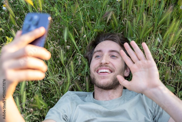Fototapeta Young man lying down enjoys a video call in a natural setting, showing a cheerful attitude