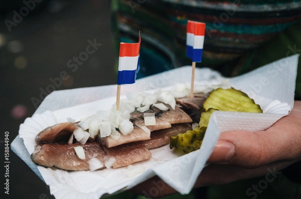Fototapeta A hand holds a traditional Dutch delicacy of herring with gherkins and onions.