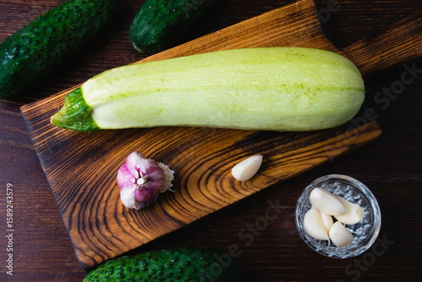 Fototapeta Cucumbers, garlic and zucchini on a cutting board, on a dark wooden background. Concept of cooking healthy food and healthy eating