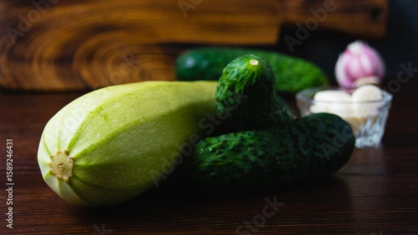Fototapeta Green vegetables on a dark background. Cucumbers, garlic and zucchini on a wooden table. Concept of cooking healthy food and healthy eating
