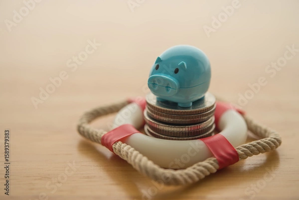 Fototapeta Blue piggy bank and stack coins in lifebuoy on wooden table background. Savings protection, financial risk management and security, savings insurance, personal and home finance and so on concept.