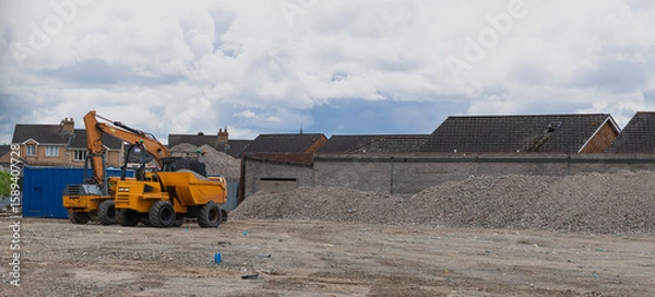 Obraz Construction site in Belfast featuring an excavator, dump truck, gravel piles, and cloudy sky—urban development and industrial activity in progress.