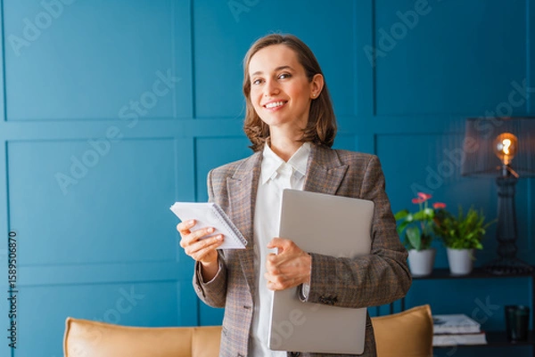 Fototapeta Beautiful woman with dark hair stands in the middle of the office with a notepad and a laptop in her hands, looking forward and smiling.