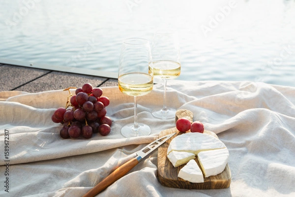 Fototapeta Two glasses of white wine with cheese and grapes on a pier at sunset.