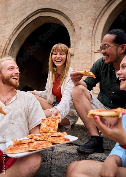 Fototapeta Vertical. Cheerful young friends enjoying delicious pizza together, laughing and sharing joyful moments while sitting on stone steps in an urban setting filled with vibrant city life