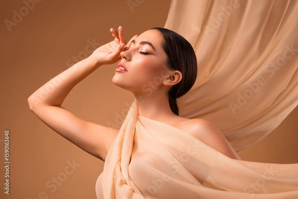 Obraz Portrait of a young brunette woman against a beige background wrapped in soft fabric with elegant expressions.
