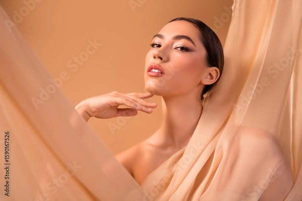 Obraz Beautiful woman with elegant makeup posing gracefully with beige fabric backdrop, emphasizing style and sensuality