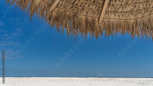 Fototapeta A fragment of a straw sun umbrella against a clear blue sky. Close-up. The upper part of the screen. The crossbars and the weave are visible. At the bottom of the frame is a white sandy beach.  