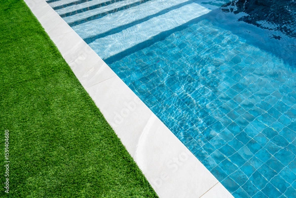 Fototapeta Swimming pool from a luxury house with white tiles around and green grass and clear transparent turquoise blue water. Real estate, property management and pool maintenance